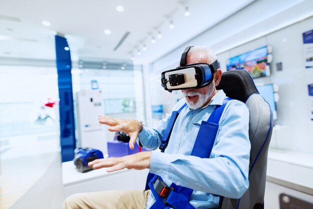 Aufgeregter bärtiger älterer Mann, der auf Stuhl sitzt und Virtual-Reality-Brille ausprobiert. Tech Store Interieur.