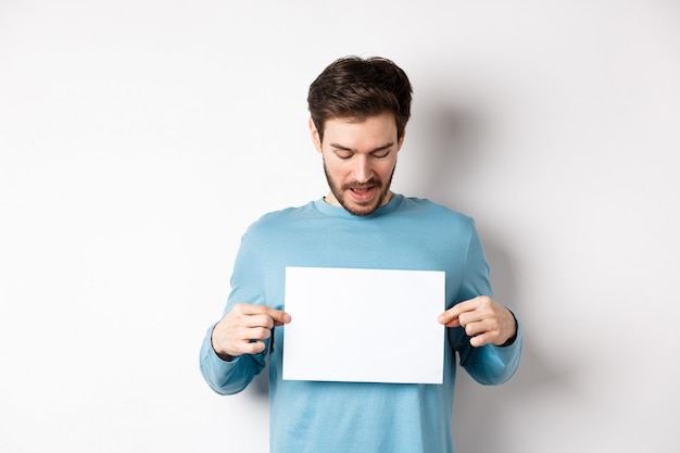 Foto aufgeregter bärtiger kerl, der fahne auf leerem blatt papier liest, logo zeigt, über weißem hintergrund stehend.