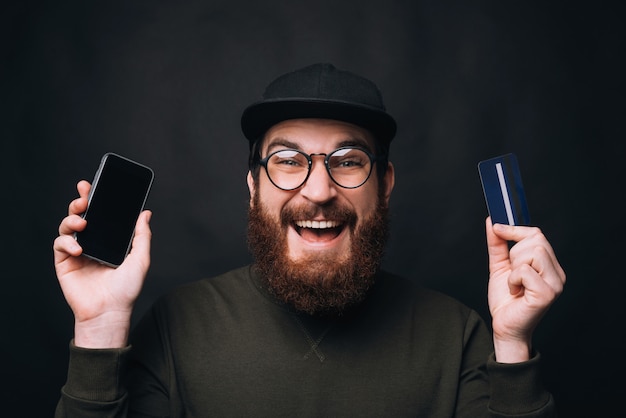 Aufgeregter bärtiger Mann, der Brille und Mütze trägt, hält eine Karte und sein Telefon.