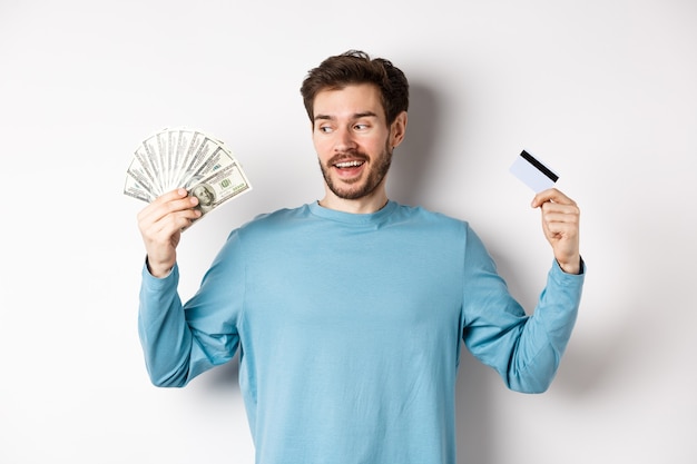 Aufgeregter bärtiger Mann, der zwischen Geld und Plastikkreditkarte wählt, Zahlung mit Bargeld oder kontaktlos, über weißem Hintergrund stehend.