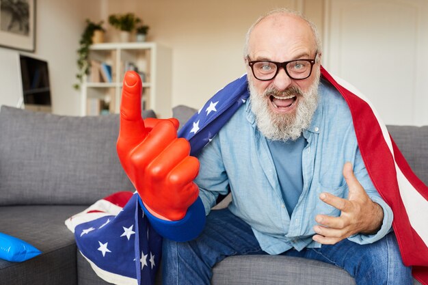 Aufgeregter bärtiger Mann in Brille mit amerikanischer Flagge, die auf Sofa sitzt und zu Hause für seine Fußballmannschaft jubelt