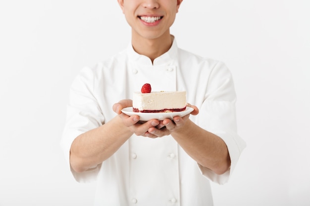 aufgeregter chinesischer Hauptmann in der weißen Kochuniform, die an der Kamera lächelt, während Platte mit leckerem Käsekuchen über weißer Wand lokalisiert hält