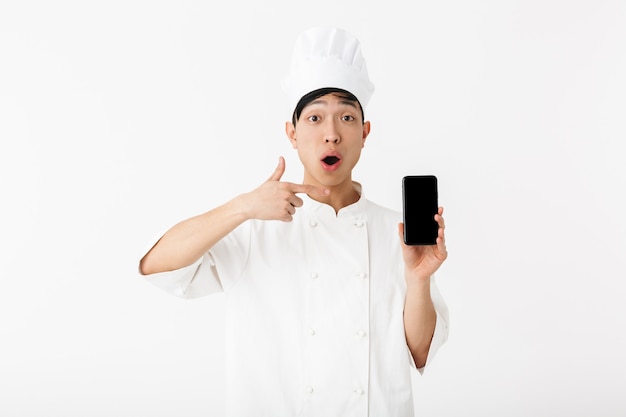 aufgeregter chinesischer Hauptmann in weißer Kochuniform und Kochmütze, die Handy lokalisiert über weißer Wand hält