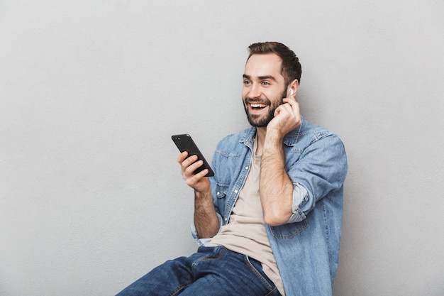 Aufgeregter fröhlicher Mann, der Hemd trägt, das über graue Wand lokalisiert wird, Kopfhörer trägt und Handy verwendet