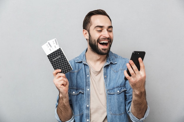 Aufgeregter fröhlicher Mann, der Hemd trägt, das über graue Wand lokalisiert wird, Pass mit Flugtickets hält, mit Handy