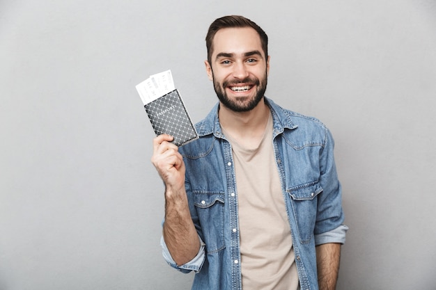 Aufgeregter fröhlicher Mann, der Hemd trägt, das über graue Wand lokalisiert wird und Pass mit Flugtickets hält