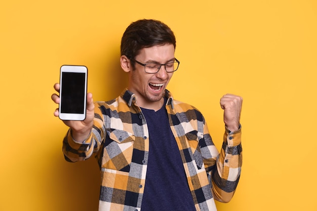 Aufgeregter froher junger Mann, der mit Smartphone über gelbem Hintergrund posiert