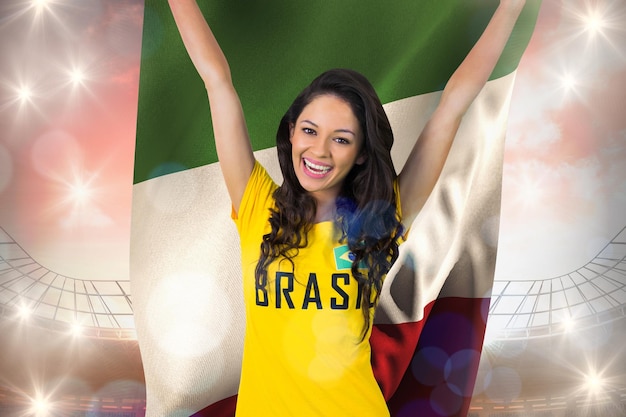 Aufgeregter fußballfan im brasil-t-shirt, das italien-flagge gegen großes fußballstadion unter bewölktem blauem himmel hält