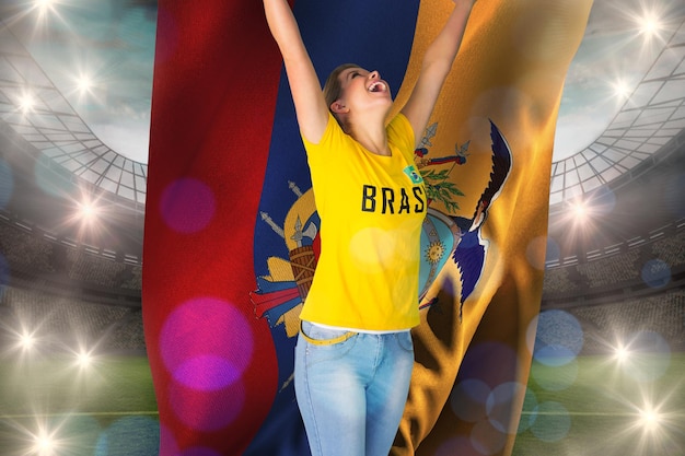 Aufgeregter fußballfan im brasilianischen t-shirt, das ecuador-flagge gegen großes fußballstadion mit lichtern hält