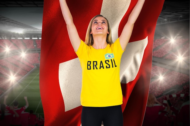 Aufgeregter Fußballfan im brasilianischen T-Shirt mit Schweizer Flagge gegen riesiges Fußballstadion mit Fans in Rot