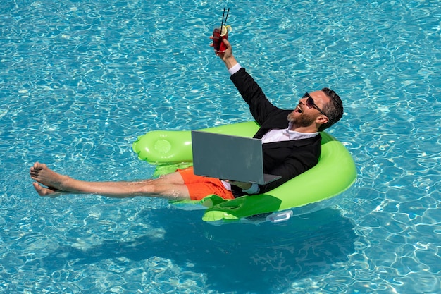 Aufgeregter Geschäftsmann, der im Anzug schwimmt, trinkt Sommercocktail und benutzt Laptop im Poolreisegeschäft