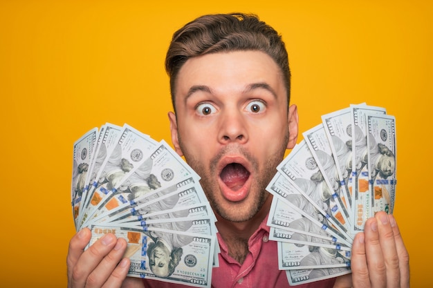 Aufgeregter, glücklicher, erfolgreicher Mann in Freizeitkleidung posiert mit Geld in den Händen, während er auf gelbem Hintergrund isoliert ist