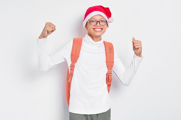 Aufgeregter hübscher junger asiatischer Student in der Weihnachtsmütze, die ein T-Shirt mit Rucksack trägt, der den Sieg lokalisiert auf weißem Hintergrund feiert