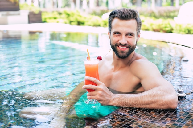 Aufgeregter hübscher moderner muskulöser Mann ruht sich im Sommerpool aus und trinkt erfrischenden Cocktail und schaut vor der Kamera.