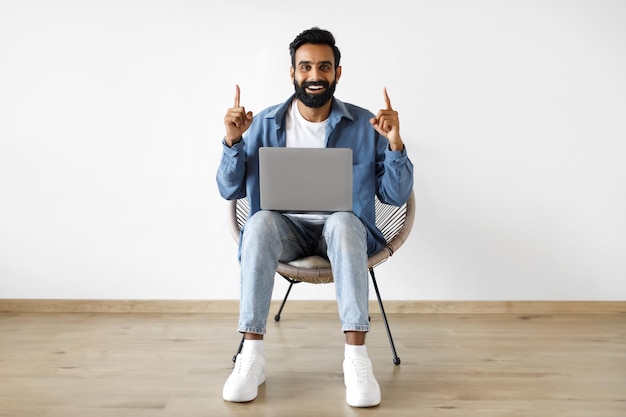 Aufgeregter indischer Mann mit Laptop, der mit den Fingern nach oben zeigt und drinnen sitzt