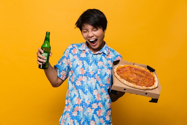 Aufgeregter junger asiatischer Mann, der isoliert über gelbem Raum steht, der Pizza hält, die Bier trinkt.