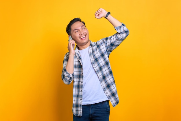 Aufgeregter junger asiatischer Mann im karierten Hemd, der am Handy spricht und großes Glück feiert, isoliert auf gelbem Hintergrund