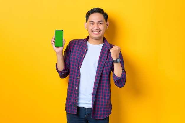 Aufgeregter junger asiatischer Mann im karierten Hemd, der ein Smartphone mit leerem Bildschirm zeigt und eine Siegergeste isoliert auf gelbem Hintergrund macht