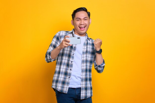 Aufgeregter junger asiatischer Mann im karierten Hemd, der Erfolg mit einem Handy feiert, das die Hände hebt, freute sich über großes Glück isoliert auf gelbem Hintergrund
