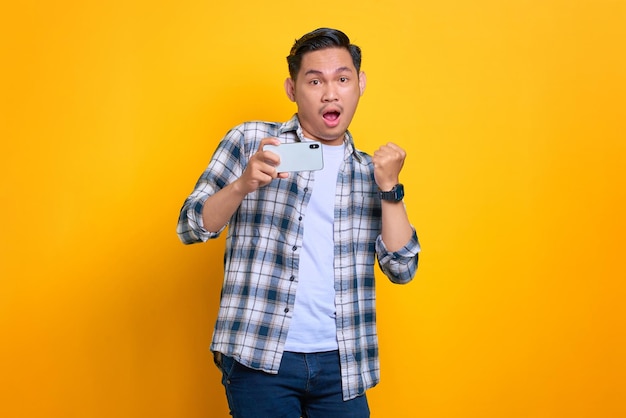 Aufgeregter junger asiatischer Mann im karierten Hemd, der Erfolg mit einem Handy feiert, das die Hände hebt, freute sich über großes Glück isoliert auf gelbem Hintergrund