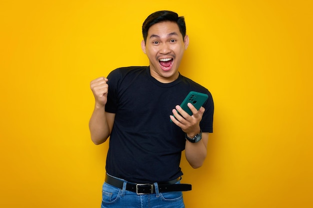 Aufgeregter junger asiatischer Mann in legerem T-Shirt mit Handy, der eine Gewinnergeste macht und gute Nachrichten isoliert auf gelbem Hintergrund erhält Lifestyle-Konzept für Menschen