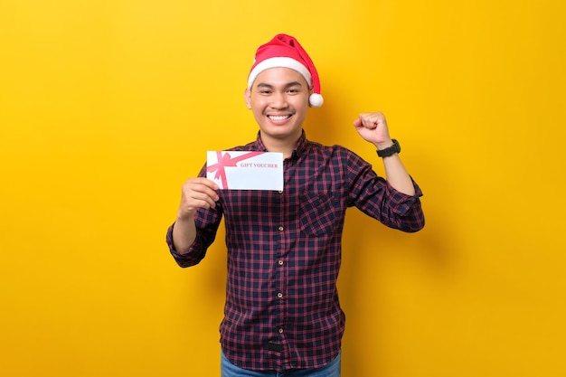 Aufgeregter junger asiatischer Mann in Weihnachtsmütze mit Geschenkgutschein, der Gewinnergeste über gelbem Studiohintergrund macht Frohes neues Jahr 2023 Feier frohes Feiertagskonzept