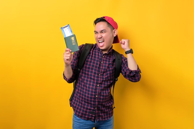 Aufgeregter junger asiatischer Mann mit Rucksack, der Reisepass und Flugtickets hält und über die Reise feiert, die über gelbem Hintergrund isoliert ist Konzept für Tourismus- und Urlaubsreisen