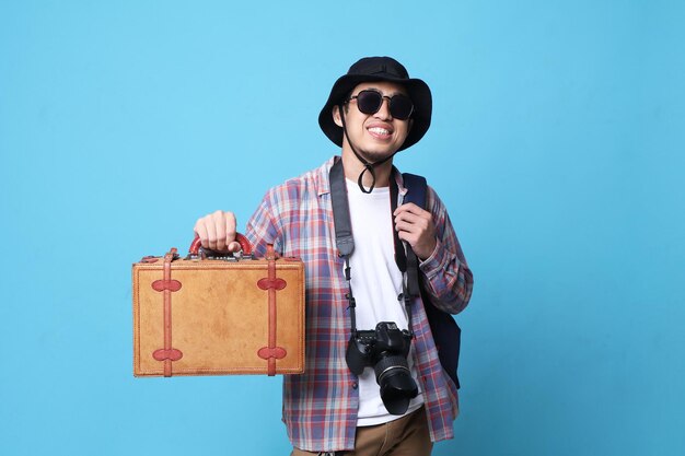 Aufgeregter junger asiatischer Mann reist ins Ausland und hält Reisetaschen isoliert über einem blauen Farbhintergrund.