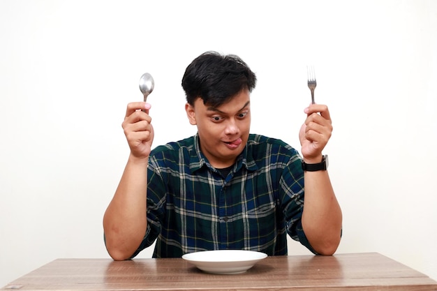 Aufgeregter junger asiatischer Mann zeigt den Daumen mit einem leeren Teller auf dem Tisch