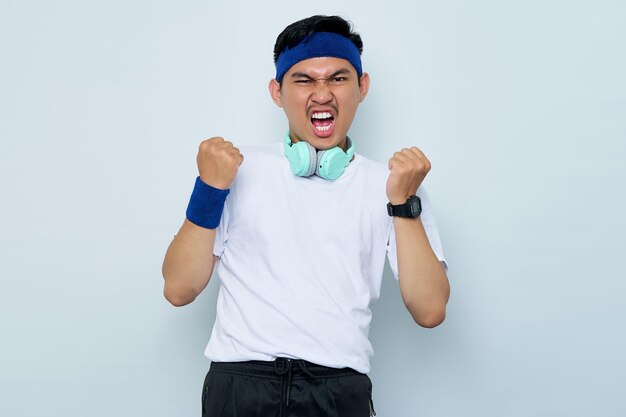 Aufgeregter junger asiatischer Sportler mit blauem Stirnband und weißem Sportbekleidungs-T-Shirt mit Kopfhörern, der den Sieg isoliert auf weißem Hintergrund feiert Workout-Sportkonzept