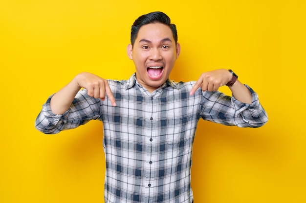Aufgeregter junger gutaussehender asiatischer Mann, der ein kariertes Hemd trägt und mit den Fingern nach unten zeigt und freien Platz für Werbung zeigt, die auf gelbem Hintergrund isoliert ist Lifestyle-Konzept für Menschen