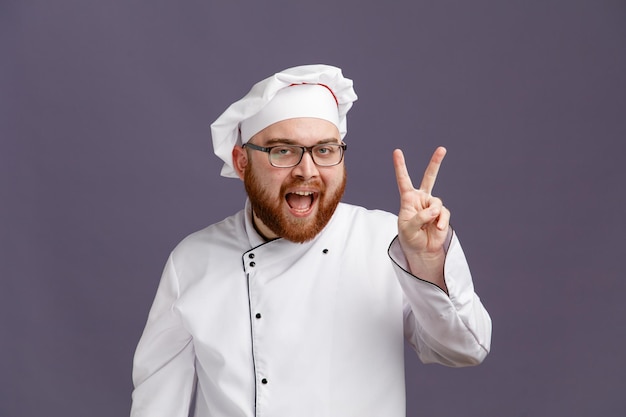 Aufgeregter junger Koch mit Brille, Uniform und Mütze, der in die Kamera blickt und das Friedenszeichen isoliert auf violettem Hintergrund zeigt