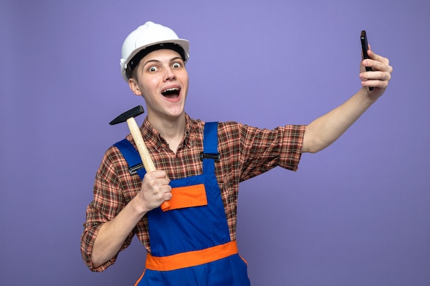 Aufgeregter junger männlicher Baumeister, der Uniform mit Hammer trägt, macht ein Selfie