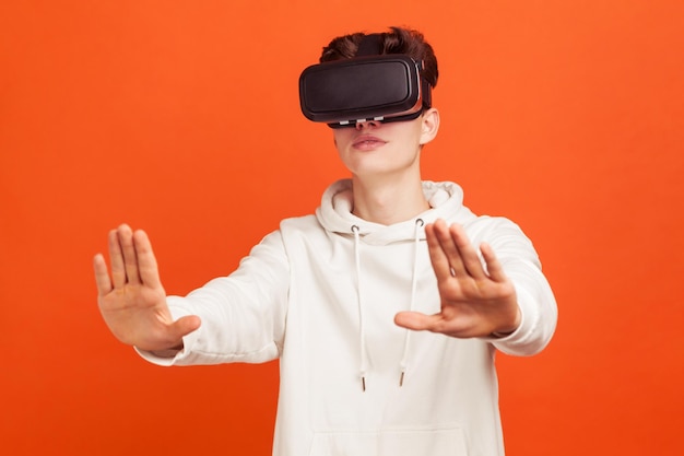 Aufgeregter junger Mann im lässigen Sweatshirt im Virtual-Reality-Headset, der eine Online-Konferenz durchführt, Kollegen, die 3D-Projekte überprüfen und diskutieren. Studioaufnahme im Innenbereich isoliert auf orangefarbenem Hintergrund