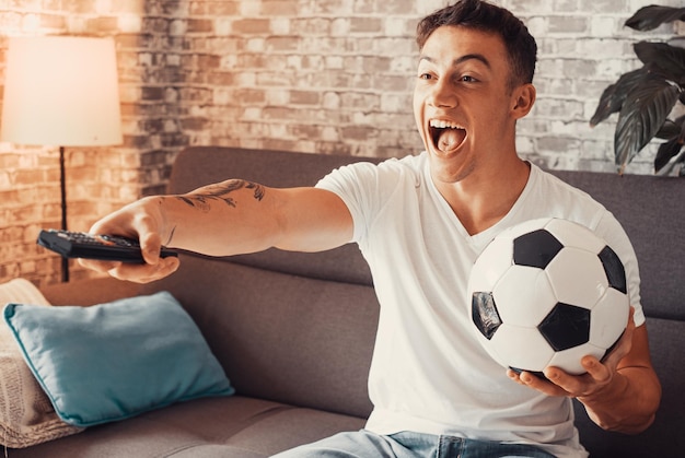 Foto aufgeregter kleiner fußballfan, der ein spiel im fernsehen sieht und einen fußball in der hand hält, während er auf einem bequemen sofa in seinem wohnzimmer sitzt