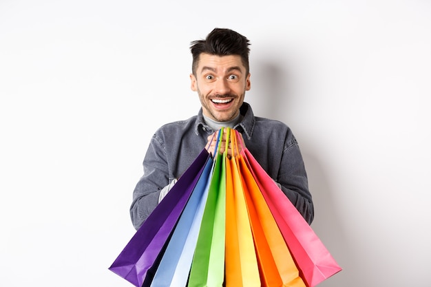 Aufgeregter lächelnder Kerl, der bunte Einkaufstaschen hält und sich mit Rabatten im Laden freut, die gegen weißen Hintergrund stehen.