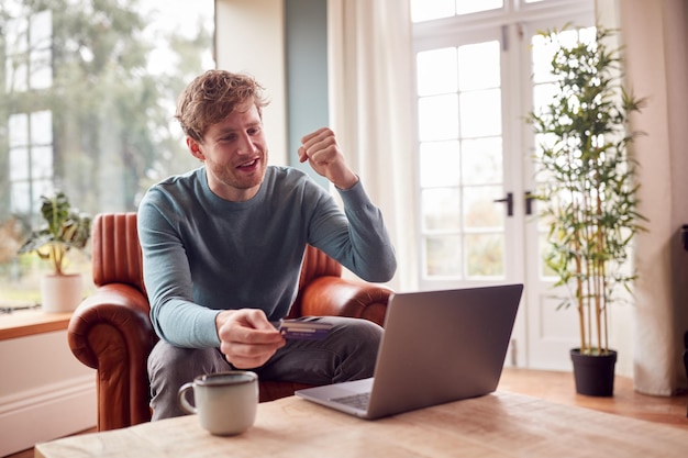 Aufgeregter Mann, der zu Hause mit Laptop auf Sessel sitzt und Online-Kauf mit Kredit- oder Debitkarte tätigt