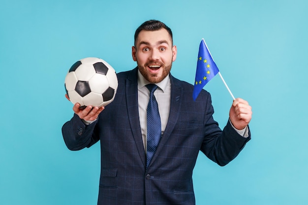 Aufgeregter Mann im Anzug im offiziellen Stil mit schwarz-weißem Fußball und EU-Flagge, der Fußball sieht und das Lieblingsteam unterstützt Indoor-Studioaufnahme isoliert auf blauem Hintergrund