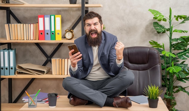 Aufgeregter Mann mit Smartphone freut sich über den Sieg und macht eine Siegesgestur, während er mit gekreuzten Beinen auf dem Büro-Schreibtisch sitzt