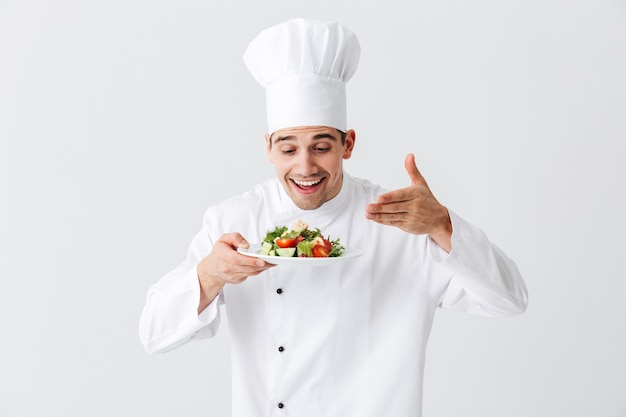 Aufgeregter Mannkochkoch, der Uniform trägt, die frischen grünen Salat auf einem Teller lokalisiert über weißer Wand zeigt
