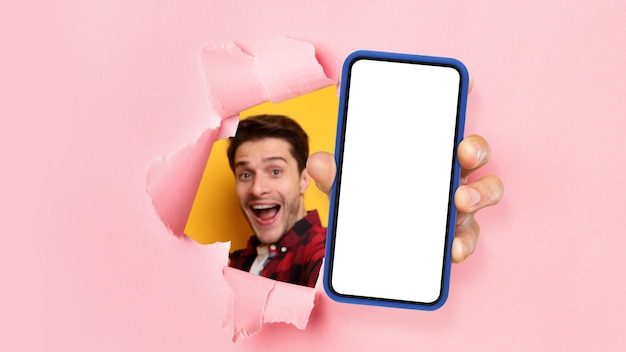 Aufgeregter Typ, der weißen, leeren Smartphone-Bildschirm durch zerrissenes Papier zeigt