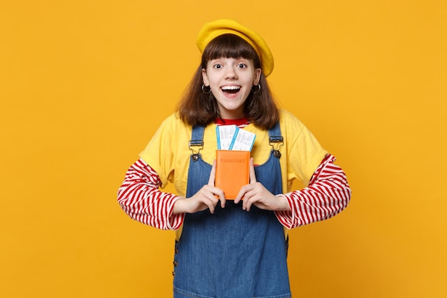 Aufgeregter überraschter Teenager in französischer Baskenmütze, Denim-Sommerkleid mit Pass-Bordkarte isoliert auf gelbem Wandhintergrund. Menschen aufrichtige Emotionen Lifestyle-Konzept. Mock-up-Kopienbereich.