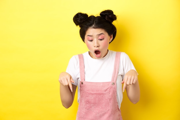 Aufgeregtes asiatisches junges Mädchen, das Werbung auscheckt, Finger nach unten zeigt und erstaunt den Kiefer fallen lässt, Promo-Angebot zeigt, gelb.