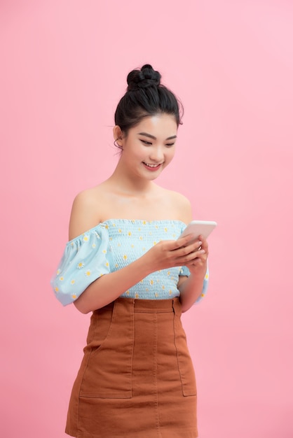 Aufgeregtes asiatisches Mädchen, das das Smartphone verwendet, das über rosa Hintergrund steht. Freiraum