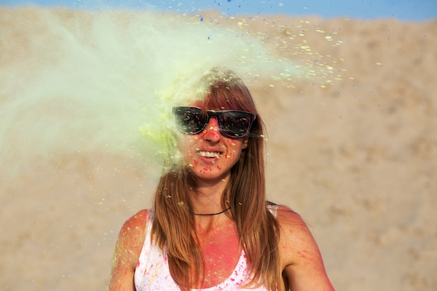 Aufgeregtes blondes Model mit Sonnenbrille, das mit grüner Holi-Farbe in der Wüste spielt