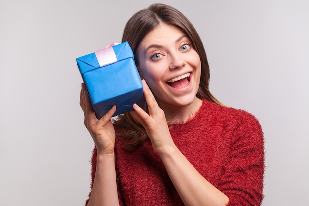 Aufgeregtes glückliches Mädchen, das Geschenkbox in der Nähe des Ohrs hält und zuhört, raten Sie, was drin ist, in Erwartung