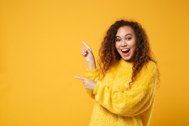 Aufgeregtes junges afrikanisch-amerikanisches Mädchen im Pelzpullover posiert isoliert auf gelb-orangefarbenem Wandhintergrund. Menschen aufrichtige Emotionen Lifestyle-Konzept. Mock-up-Kopienbereich. Zeigefinger zur Seite zeigen.