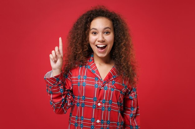 Aufgeregtes junges afrikanisch-amerikanisches Mädchen in Pyjama-Homewear, das zu Hause isoliert auf rotem Hintergrund ruht. Entspannen Sie sich bei guter Laune Lifestyle-Konzept. Mock-up-Kopienbereich. Zeigefinger hochhalten mit toller neuer Idee.