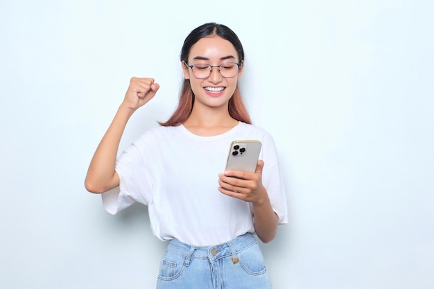 Aufgeregtes junges asiatisches Mädchen im weißen T-Shirt mit Smartphone und Gewinnergeste isoliert auf weißem Hintergrund