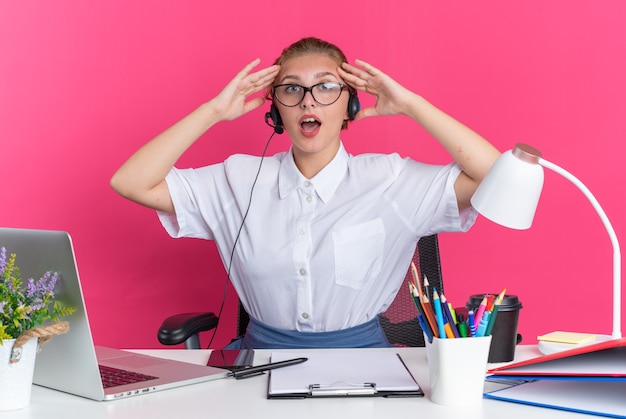 Aufgeregtes junges blondes Call-Center-Mädchen mit Headset und Brille, das am Schreibtisch mit Arbeitswerkzeugen sitzt und die Hände auf dem Kopf hält und die Kamera isoliert auf rosa Wand betrachtet